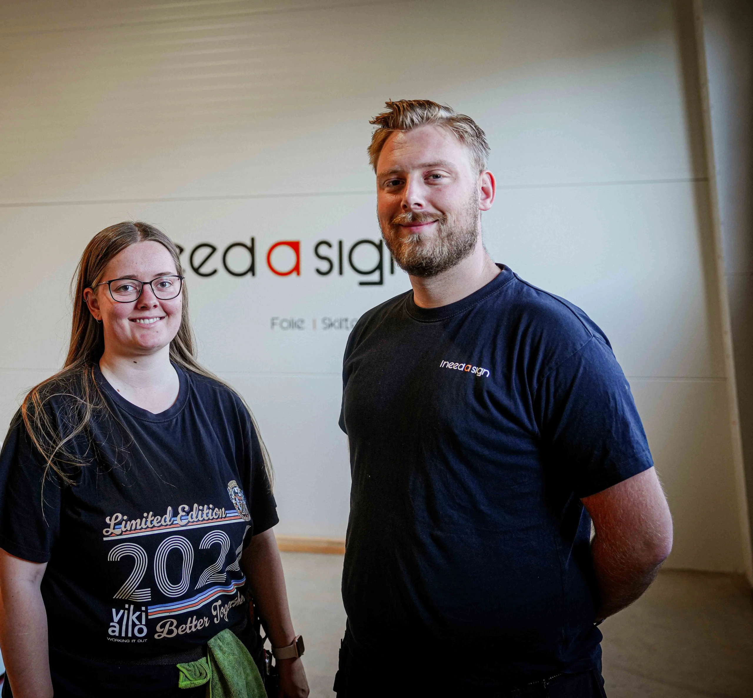 Patrick og Alexandra står side om side foran logoet på væggen i værkstedet