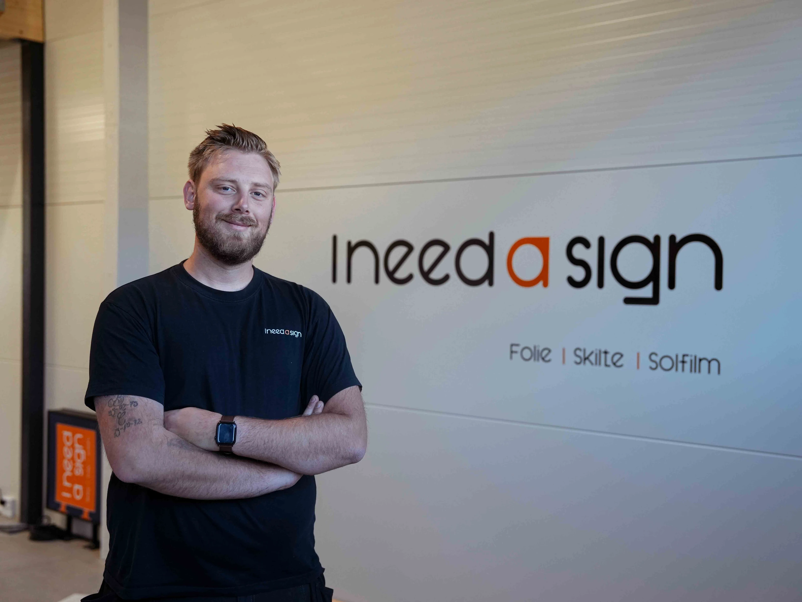 Patrick, indehaver af I need a sign, med armene over kors foran logoet på væggen i værkstedet